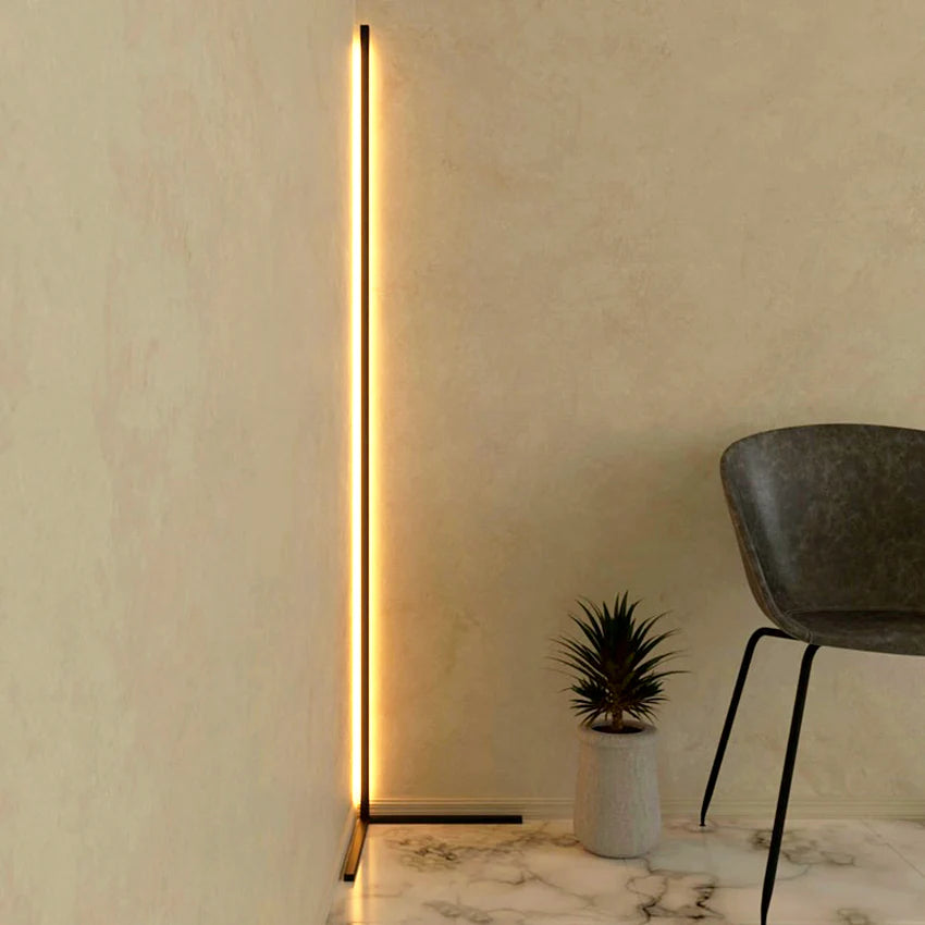 A view of the Enlights LED vertical strip corner lamp on black a V stand on high brightness against a white wall next to a black chair and a small plant. 