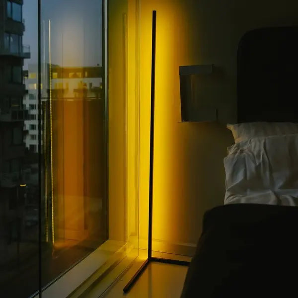 An Enlights LED vertical strip lamp in the corner of a high-rise apartment next to a bed. The Enlight glows with a bright gold illumination. 