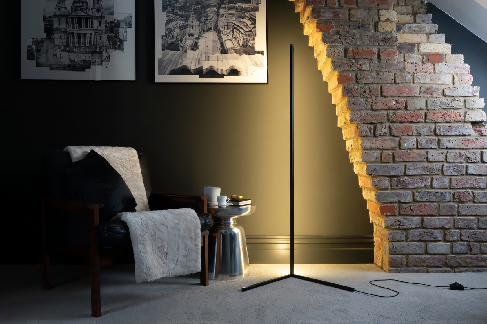An Enlight LED vertical strip light on a V stand lighting a corner in a home with a brick wall accent, a cozy chair, chic side table, and art on a earth tone wall. 
