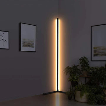 A front of view of the Enlights LED vertical strip corner lamp on black a V stand on high brightness against a white wall with plants and art. 